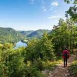 S'évader en plein cœur du tourisme à Hauteville-Lompnes Femme en veste rouge marchant sur un sentier forestier. Vue sur une rivière sinueuse et des montagnes boisées sous un ciel bleu.