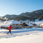 Skieur de fond en rouge sur une piste de Corrençon en Vercors, avec des chalets enneigés et montagnes boisées sous un ciel clair.