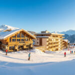 Trouver le logement à La Rosière idéal pour votre séjour Vue panoramique de la station de ski La Rosière avec skieurs, chalets en bois, résidences modernes et montagnes enneigées sous ciel bleu.