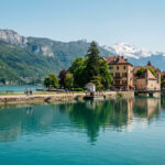 Canal turquoise d'Annecy avec ses rives, bâtiments colorés, promeneurs et l'arrière-plan des montagnes alpines et enneigées.