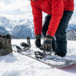 Gros plan sur une personne ajustant son snowboard et ses fixations, bottes et casque à proximité, avec un fond de montagnes enneigées.