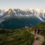 Deux randonneurs marchent sur un sentier rocailleux vers un refuge de montagne, avec les sommets enneigés du Mont Blanc illuminés par le soleil levant en arrière-plan.