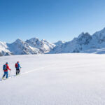 Deux skieurs de randonnée progressent sur un plateau enneigé. Des montagnes imposantes dominent l'horizon sous un ciel bleu limpide.