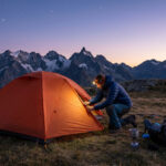 Réussir son premier bivouac : tout ce qu'il faut savoir Randonneur préparant sa tente orange au coucher du soleil face à des sommets enneigés.