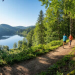Commencer le trail dans les Vosges : 3 boucles faciles Un homme et une femme courent sur un sentier en forêt surplombant un lac paisible dans les Vosges par une journée ensoleillée.