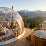 Séjour glamping dans les Alpes : le luxe en pleine nature Un dôme géodésique transparent avec un lit et un jacuzzi en bois sur une terrasse surplombant des montagnes enneigées.