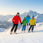 Les stations de Savoie idéales pour skier en famille Une famille souriante skie ensemble sur une piste ensoleillée avec les montagnes enneigées en arrière-plan.