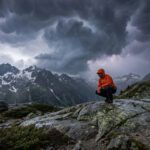 Survivre à un orage en montagne : les gestes qui sauvent Randonneur en veste orange accroupi sur un rocher face à un orage menaçant au-dessus des Alpes.