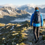 Un randonneur portant une veste bleue et un sac à dos marche sur un sentier face à un panorama de sommets enneigés.