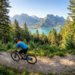 Parcourir Annecy en VTT : 5 circuits incontournables Vététiste en maillot bleu roulant sur un sentier forestier avec vue panoramique sur le lac d'Annecy et les montagnes.