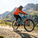 Un vététiste en équipement orange sur un VTT électrique bleu descendant un sentier rocheux en montagne.